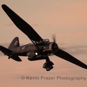 May include: A black and red vintage airplane in flight against a pink and orange sunset sky. The airplane has a propeller and a visible cockpit.