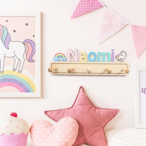 Puede incluir: Un perchero de pared de madera con el nombre Naomi en letras pastel, un peque&ntilde;o arco&iacute;ris y una figura de unicornio. Se muestra entre dos l&aacute;minas enmarcadas con un unicornio y el texto unicorn vibes, con banderines rosas arriba.