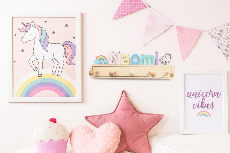Puede incluir: Un perchero de pared de madera con el nombre Naomi en letras pastel, un peque&ntilde;o arco&iacute;ris y una figura de unicornio. Se muestra entre dos l&aacute;minas enmarcadas con un unicornio y el texto unicorn vibes, con banderines rosas arriba.