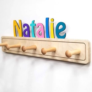 Op de afbeelding: Een houten kapstok met zes houten haken en een naambord boven de haken. Het naambord is gemaakt van kleurrijke letters die "Natalie" spellen.