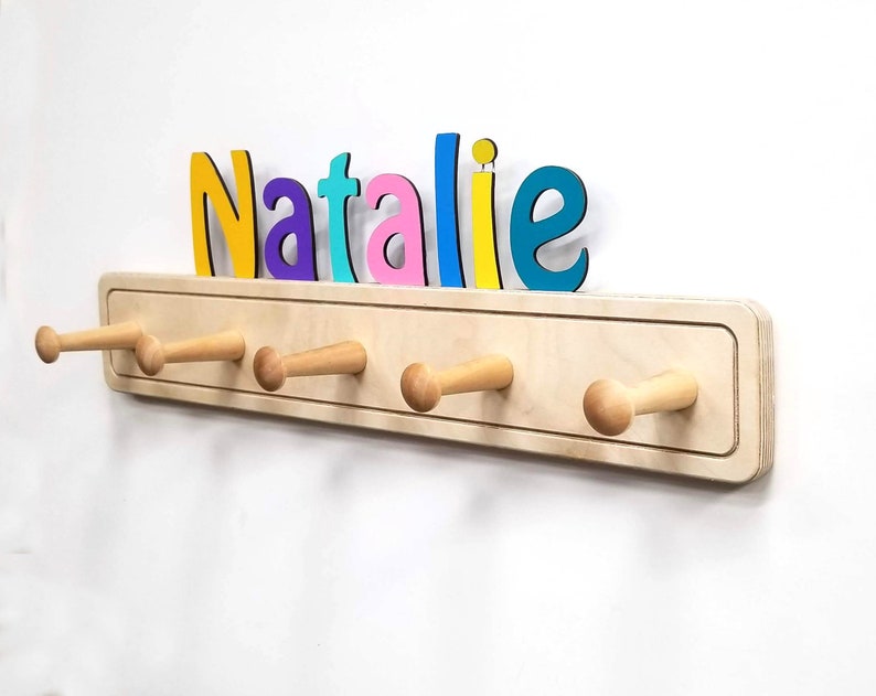 Op de afbeelding: Een houten kapstok met zes houten haken en een naambord boven de haken. Het naambord is gemaakt van kleurrijke letters die "Natalie" spellen.