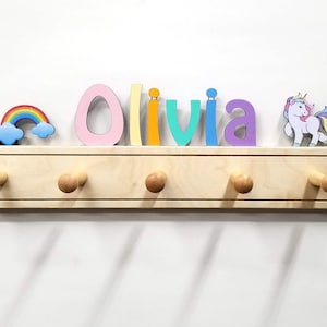 Puede incluir: Un perchero de pared de madera clara con cinco ganchos redondeados. La parte superior est&aacute; decorada con el nombre Olivia en letras multicolores, un arco&iacute;ris con nubes azules a la izquierda y un unicornio blanco con crines de colores a la derecha.