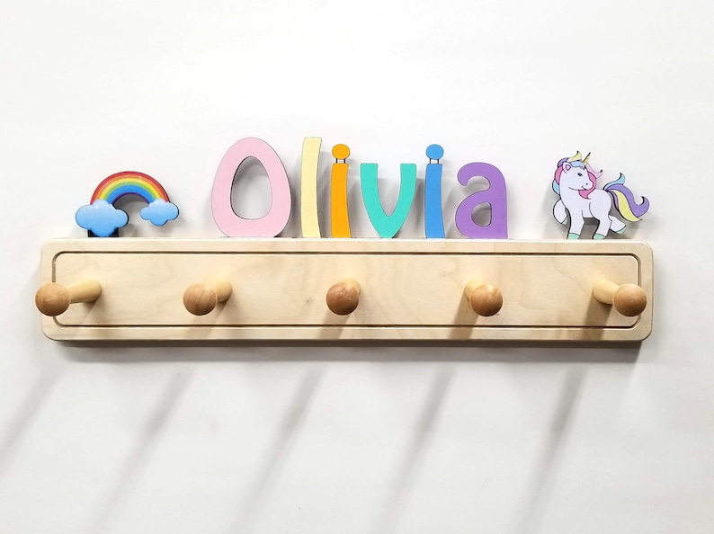 Puede incluir: Un perchero de pared de madera clara con cinco ganchos redondeados. La parte superior est&aacute; decorada con el nombre Olivia en letras multicolores, un arco&iacute;ris con nubes azules a la izquierda y un unicornio blanco con crines de colores a la derecha.