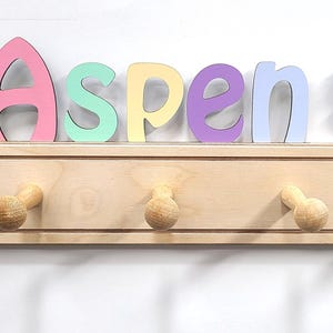 Puede incluir: Un perchero de pared de madera clara con cinco ganchos redondeados. Sobre el perchero, el nombre Aspen est&aacute; escrito con letras coloridas, flanqueado por una figura de princesa a la izquierda y una figura de unicornio a la derecha.
