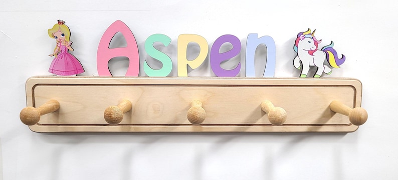 Puede incluir: Un perchero de pared de madera clara con cinco ganchos redondeados. Sobre el perchero, el nombre Aspen est&aacute; escrito con letras coloridas, flanqueado por una figura de princesa a la izquierda y una figura de unicornio a la derecha.