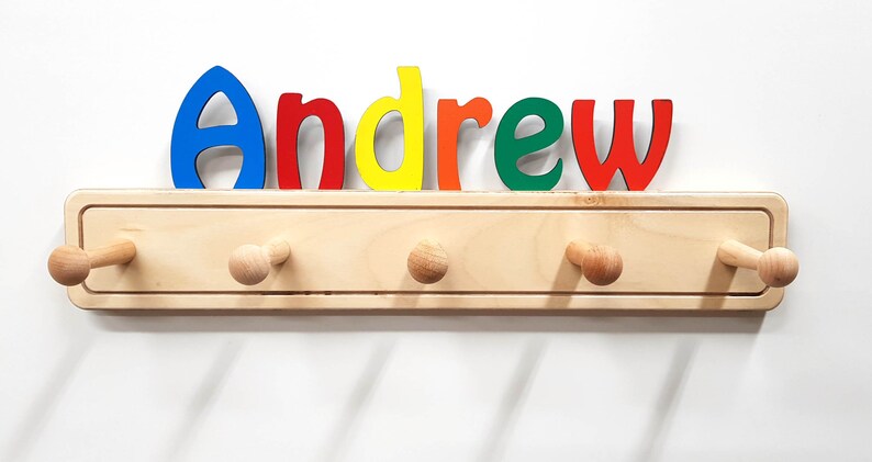 Op de afbeelding: Een houten kapstok met vijf haken en de naam "Andrew" gespeld in kleurrijke letters boven de haken.