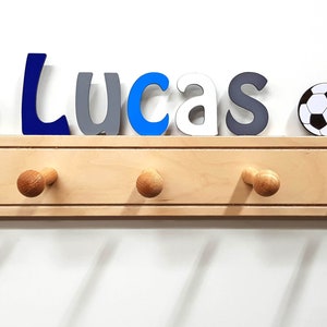Puede incluir: Un perchero de pared de madera clara con cinco ganchos redondeados. Sobre el perchero, el nombre Lucas est&aacute; escrito con letras individuales en azul oscuro, gris, azul claro y blanco, flanqueado por dos balones de f&uacute;tbol en blanco y negro.