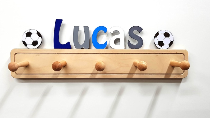 Puede incluir: Un perchero de pared de madera clara con cinco ganchos redondeados. Sobre el perchero, el nombre Lucas est&aacute; escrito con letras individuales en azul oscuro, gris, azul claro y blanco, flanqueado por dos balones de f&uacute;tbol en blanco y negro.