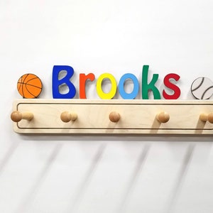 Puede incluir: Un perchero de pared de madera clara con cinco ganchos y el nombre Brooks en letras coloridas. Un icono de baloncesto est&aacute; a la izquierda y un icono de b&eacute;isbol a la derecha. Las letras son de color azul, naranja, amarillo, azul claro, verde y rojo.