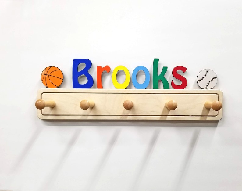 Puede incluir: Un perchero de pared de madera clara con cinco ganchos y el nombre Brooks en letras coloridas. Un icono de baloncesto est&aacute; a la izquierda y un icono de b&eacute;isbol a la derecha. Las letras son de color azul, naranja, amarillo, azul claro, verde y rojo.