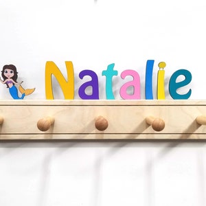 Puede incluir: Un perchero de pared de madera clara con cinco ganchos redondeados y una figura decorativa de una sirena. El nombre Natalie aparece en grandes letras de madera multicolor en amarillo, morado, azul claro, rosa y turquesa sobre el perchero.