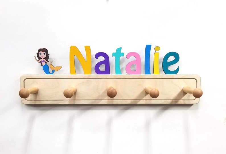 Puede incluir: Un perchero de pared de madera clara con cinco ganchos redondeados y una figura decorativa de una sirena. El nombre Natalie aparece en grandes letras de madera multicolor en amarillo, morado, azul claro, rosa y turquesa sobre el perchero.