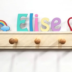 Puede incluir: Un perchero de pared de madera clara con cinco ganchos redondeados. Sobre el perchero, el nombre Elise est&aacute; escrito con letras individuales de colores, acompa&ntilde;ado de un peque&ntilde;o icono de arco&iacute;ris a la izquierda y dos corazones rojos.