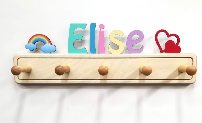 Puede incluir: Un perchero de pared de madera clara con cinco ganchos redondeados. Sobre el perchero, el nombre Elise est&aacute; escrito con letras individuales de colores, acompa&ntilde;ado de un peque&ntilde;o icono de arco&iacute;ris a la izquierda y dos corazones rojos.