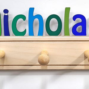 Puede incluir: Un perchero de pared de madera clara con cinco ganchos redondeados. El nombre Nicholas est&aacute; escrito en grandes letras de madera multicolor sobre el soporte. Una locomotora colorida se sit&uacute;a a la izquierda y un cami&oacute;n volquete amarillo a la derecha.