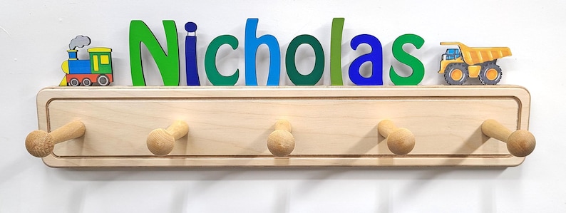Puede incluir: Un perchero de pared de madera clara con cinco ganchos redondeados. El nombre Nicholas est&aacute; escrito en grandes letras de madera multicolor sobre el soporte. Una locomotora colorida se sit&uacute;a a la izquierda y un cami&oacute;n volquete amarillo a la derecha.