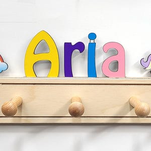 Puede incluir: Un perchero de pared de madera clara con cinco ganchos redondeados. La parte superior est&aacute; decorada con el nombre Aria en letras coloridas, un arco&iacute;ris con nubes a la izquierda y un unicornio blanco con crin morada y amarilla a la derecha.