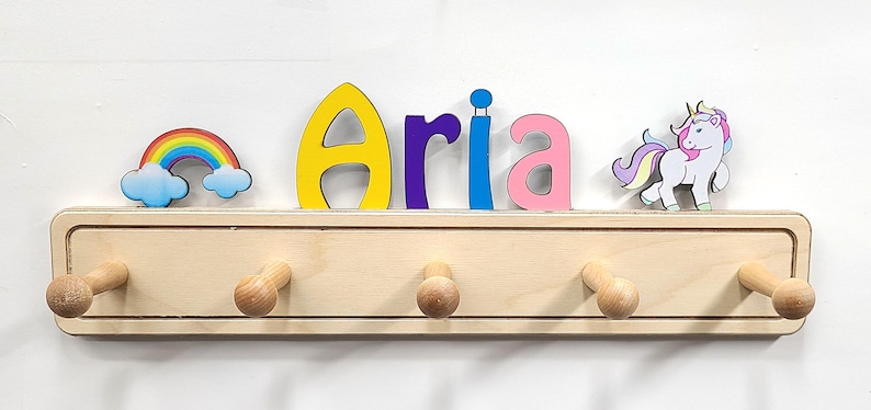 Puede incluir: Un perchero de pared de madera clara con cinco ganchos redondeados. La parte superior est&aacute; decorada con el nombre Aria en letras coloridas, un arco&iacute;ris con nubes a la izquierda y un unicornio blanco con crin morada y amarilla a la derecha.