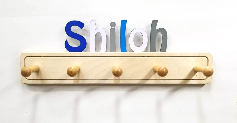 Op de afbeelding: Een houten kapstok met vijf haken en de naam "Shiloh" gespeld in kleurrijke letters boven de haken.