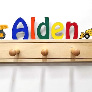 Puede incluir: Un perchero de pared de madera clara con cinco ganchos redondeados. Sobre el perchero, el nombre Alden aparece en letras grandes y multicolores, flanqueado por un cami&oacute;n volquete amarillo a la izquierda y un tractor amarillo a la derecha.