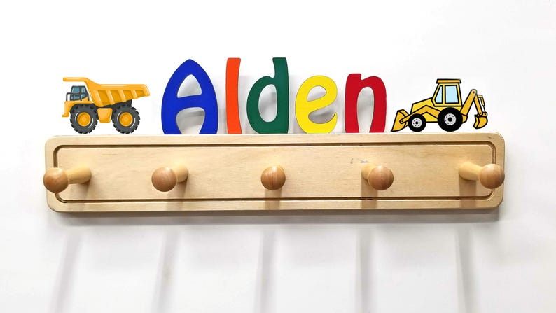 Puede incluir: Un perchero de pared de madera clara con cinco ganchos redondeados. Sobre el perchero, el nombre Alden aparece en letras grandes y multicolores, flanqueado por un cami&oacute;n volquete amarillo a la izquierda y un tractor amarillo a la derecha.