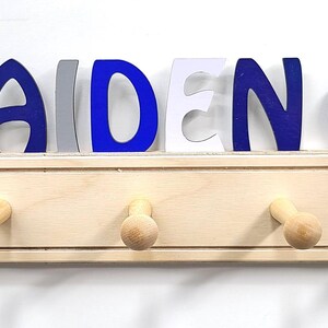 Puede incluir: Un perchero de pared de madera clara con cinco ganchos, coronado con el nombre AIDEN en letras azules, grises y blancas. Un icono de bal&oacute;n de f&uacute;tbol americano est&aacute; a la izquierda y un icono de pelota de b&eacute;isbol a la derecha del nombre, sobre una base.