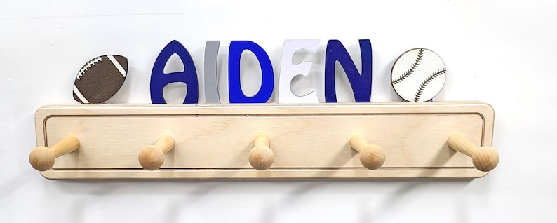 Puede incluir: Un perchero de pared de madera clara con cinco ganchos, coronado con el nombre AIDEN en letras azules, grises y blancas. Un icono de bal&oacute;n de f&uacute;tbol americano est&aacute; a la izquierda y un icono de pelota de b&eacute;isbol a la derecha del nombre, sobre una base.