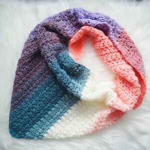 May include: A handmade crochet shawl in a gradient of colors, including purple, blue, white, and peach. The shawl is made with a textured stitch and is draped on a white, fluffy surface. The overall theme is cozy and handmade.