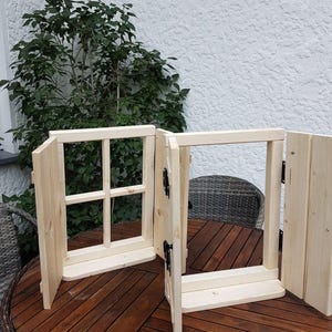 Könnte beinhalten: Zwei unfertige Holzfensterrahmen mit Fensterläden, auf einem Holztisch. Ein Fenster hat ein kreuzförmiges Gitter, während das andere offen ist. Die Fensterläden sind mit schwarzen Scharnieren befestigt. Der Hintergrund umfasst Grün und eine weiße Wand.