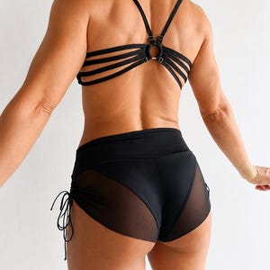 May include: A black two-piece set consisting of a strappy bra top with a central metal ring connector and matching high-waisted shorts. The shorts feature solid black panels with sheer mesh side inserts and adjustable side drawstring ties.