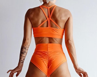 May include: A woman wearing a bright orange sports bra and matching high-waisted shorts. The outfit features a criss-cross design on the bra and a ruched detail on the shorts.