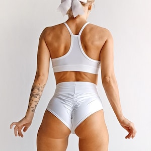 Puede incluir: Un sujetador deportivo blanco y unos pantalones cortos de cintura alta a juego con un diseño fruncido. La modelo lleva el atuendo y está de pie de espaldas a la cámara.