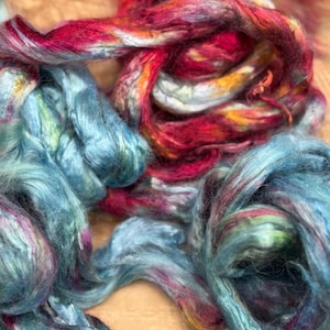 May include: Close-up of colorful fiber bundles. The fibers are a mix of blues, reds, and yellows, creating a vibrant and textured appearance. The fibers appear soft and fluffy, suggesting they are suitable for crafts like spinning or felting.