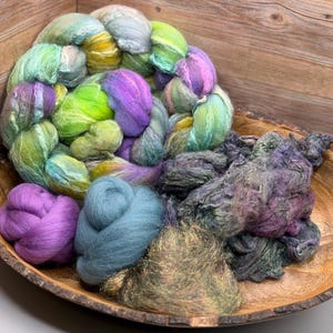 May include: A wooden bowl filled with colorful hand-dyed yarn. The yarn is a mix of green, purple, blue, and yellow. The bowl is made of wood and has a natural edge. The yarn is arranged in a variety of textures and forms, perfect for fiber art projects.