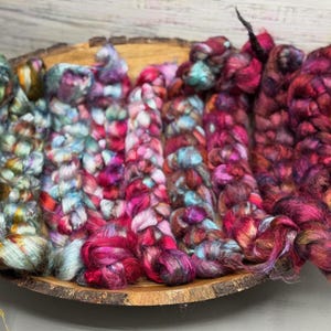May include: A wooden bowl holds several colorful braids of hand-dyed yarn. The yarn is a mix of blues, greens, pinks, and reds. The braids are thick and textured, with a variety of colors and tones. The bowl is made of wood and has a natural edge.