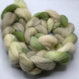 May include: A close-up of a braided roving in shades of green, beige, and cream. The fiber is soft and fluffy, with a gradient of colors. The braid is arranged in a circular shape, perfect for fiber art projects.