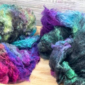 Puede incluir: Dos montones de coloridas fibras artesanales en tonos turquesa, morado, rosa y verde. La textura suave y esponjosa sugiere su idoneidad para proyectos de hilado, fieltro o tejido. Presentado sobre una superficie de madera.