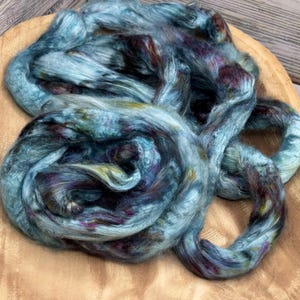 May include: A skein of hand-dyed fiber in shades of blue, purple, and green rests in a wooden bowl. The fiber is soft and fluffy, with a subtle sheen. Ideal for spinning, felting, or other fiber arts projects.