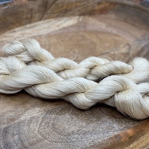 May include: A close-up of a bundle of off-white yarn, likely silk, displayed on a wooden surface.