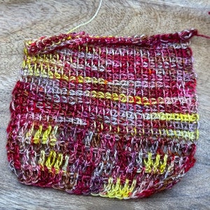 May include: A small crocheted square in shades of red, yellow, and white. The square is made with a variety of stitches and has a textured appearance.