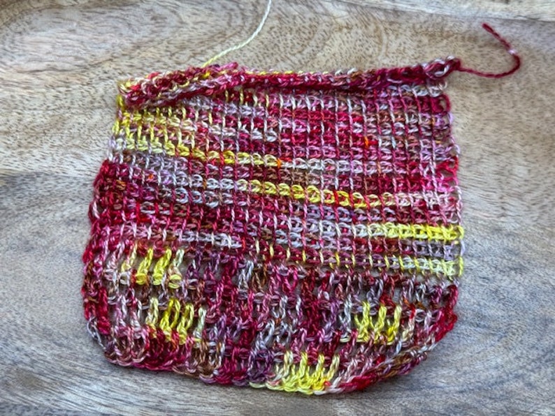 May include: A small crocheted square in shades of red, yellow, and white. The square is made with a variety of stitches and has a textured appearance.