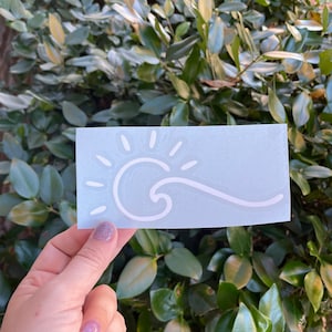 May include: White vinyl sticker with a sun and wave design on a light blue background.