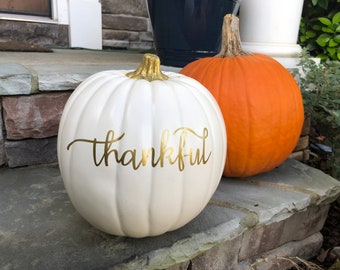 May include: A white pumpkin with the word "thankful" written in gold lettering. The pumpkin is sitting on a stone step next to an orange pumpkin.