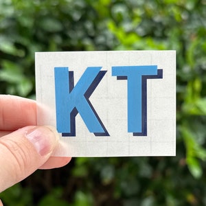 May include: A white sticker with the letters "KT" in blue with a black outline.