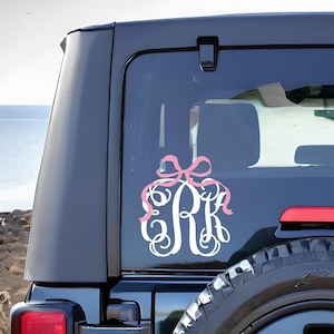 May include: A white monogram decal with the letters GRK, adorned with a pink bow, is affixed to the rear window of a black vehicle. The decal is a decorative element, adding a personalized touch to the car.