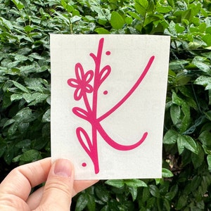 May include: A pink vinyl decal featuring a stylized letter K with a floral design. The letter is outlined in pink and has a small flower and leaves.