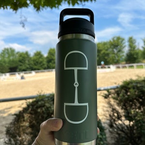 May include: A green stainless steel water bottle with a black handle and a white equestrian bit design. The bottle is held in a hand.