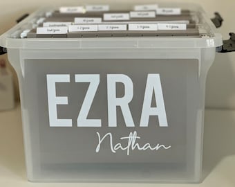 Op de afbeelding: Een doorzichtige plastic opbergdoos met een grijs label waarop "EZRA Nathan" staat. De doos bevat ordners met labels die "Kindergarten", "First Grade", "1-2 Years", "2-3 Years", "3-4 Years" en "4-5 Years" zeggen.