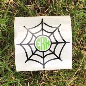 Puede incluir: Una pegatina de vinilo blanco con un diseño de telaraña negra y un monograma verde en el centro. El monograma dice "ERK".