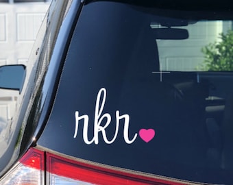 May include: A white vinyl decal with the word "rkr" in cursive script and a pink heart. The decal is on the back window of a black car.